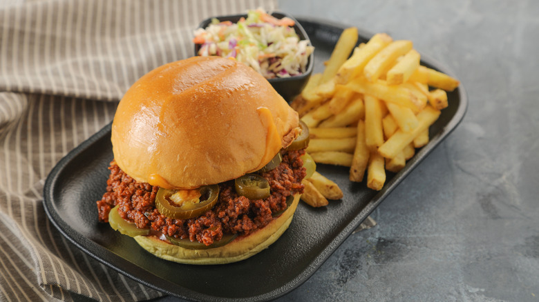A sloppy joe sandwich with jalapenos on it next to french fries and coleslaw on a black plate.