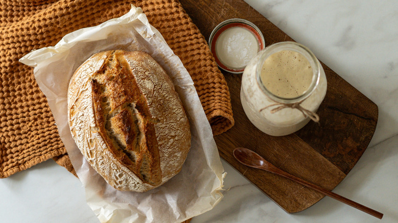 A loaf of sourdough bread on paper with a mason jar of sourdough starter nearby.