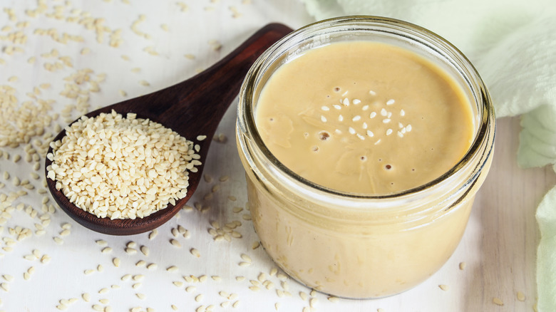 A mason jar of tahini next to a wooden spoon that is holding sesame seeds.