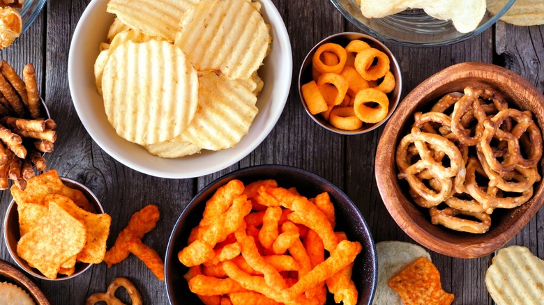 Potato chips, pretzels, tortilla chips, and Cheetos in bowls spread out on a table.