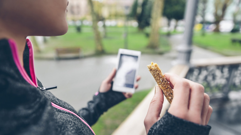 Person holding phone in one hand and granola bar in the other