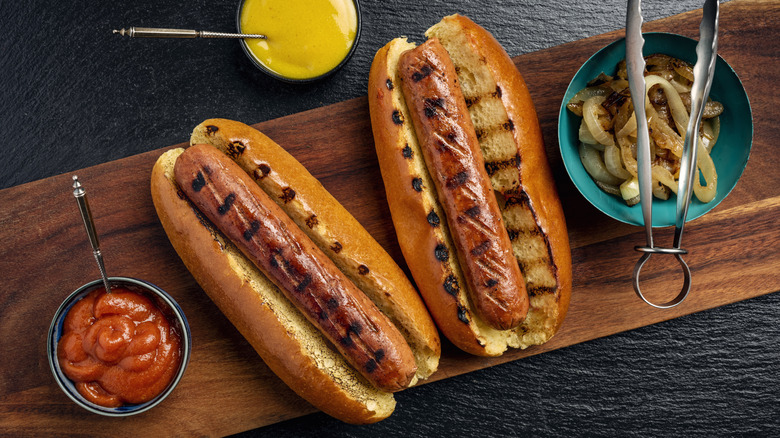 Two hot dogs with grill marks in buns surrounded by various toppings