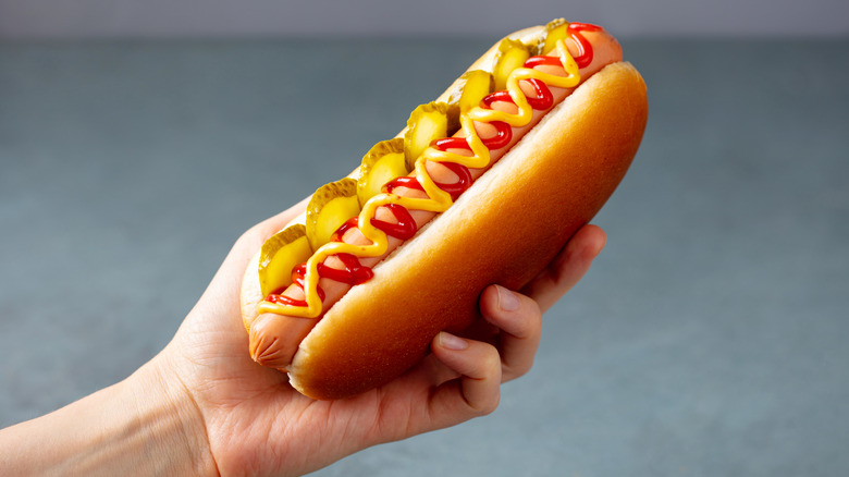 A hand holding a hot dog in a bun topped with ketchup, pickles, and mustard