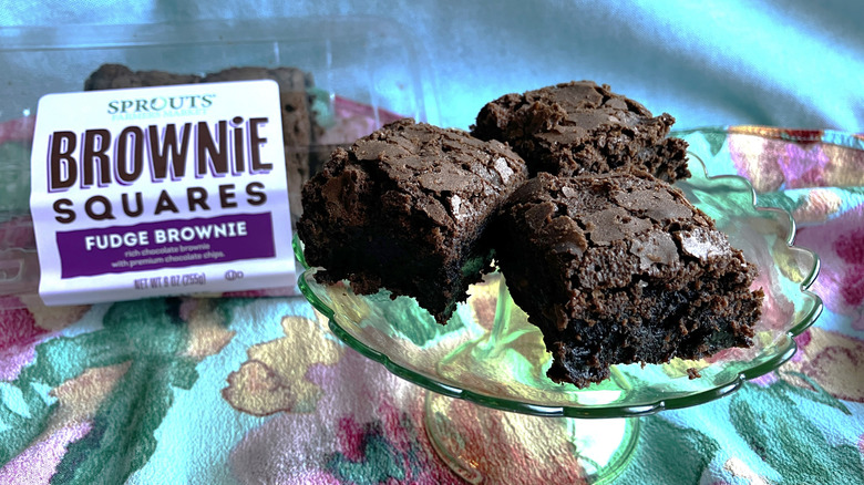 Three gooey brownies are displayed on a mini stand and the plastic container of brownies from Sprouts Farmer's Market can be seen in the background.
