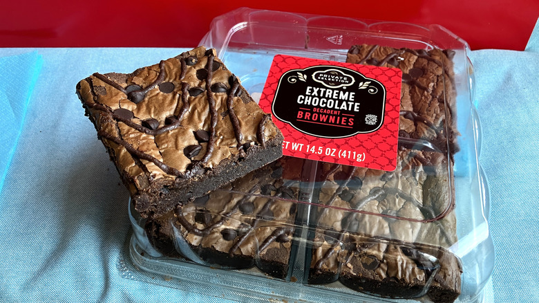 A square brownie with squiggles of chocolate and mini chocolate chips sits on top of a plastic box containing three more brownies.