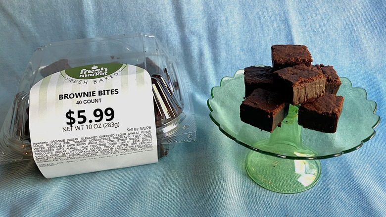 A plastic container of Fresh Market brownies sits next to a mini cake stand displaying brownie bites.