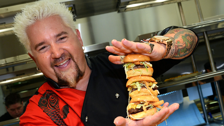 Guy Fieri holding three burgers together while smiling with his mouth open in a kitchen.