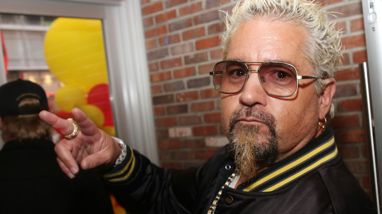 Guy Fieri wearing sunglasses entering a brick building with his pointer and middle finger up