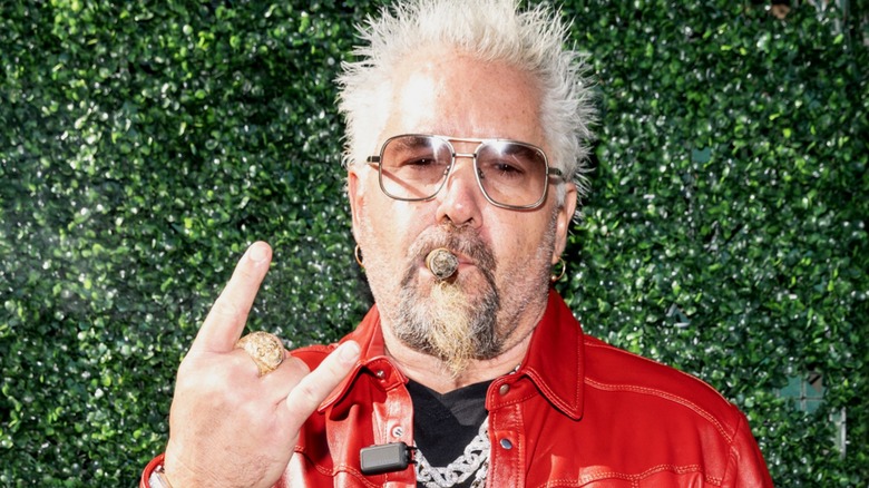 Guy Fieri posing for a photograph in front of a hedge with the devil horns up and a cigar in his mouth.