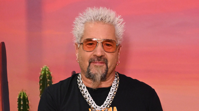 Guy Fieri wearing a chain and sunglasses holding a can in front of a painted background.