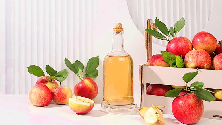 A glass container of apple juice with a bunch of apples on table and in pink slatted crate