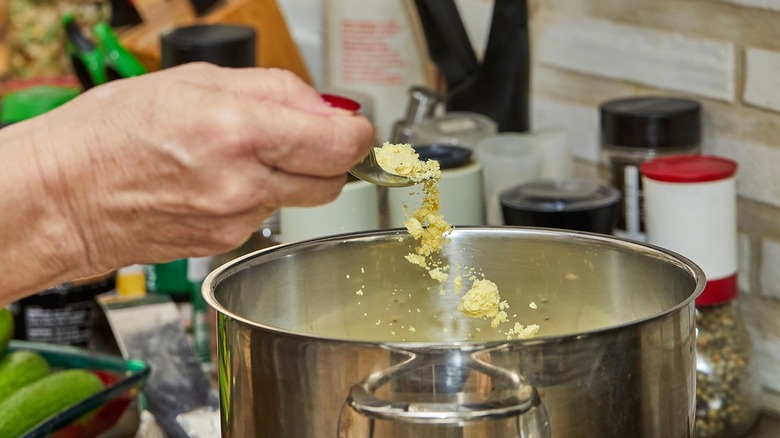 Person tossing chicken broth powder into pot with spoon on gas stove