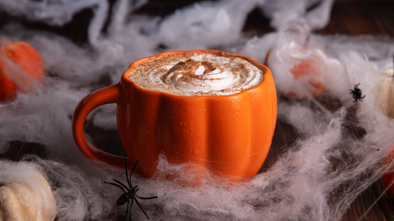 Latte in a pumpkin-shaped mug decorated with whipped cream and cinnamon surrounded by fake spiderwebs