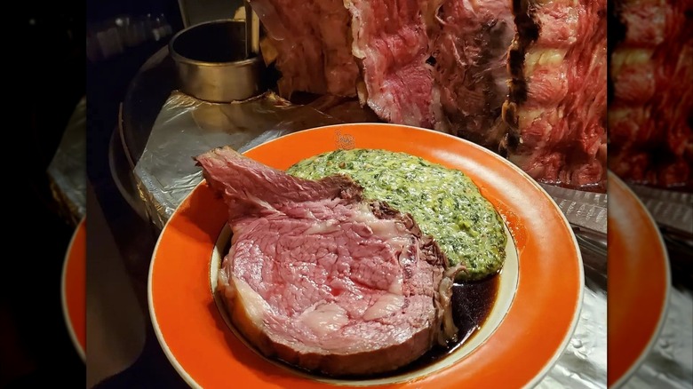 Prime rib served on House of Prime Rib's iconic orange-ringed plate with creamed spinach and a line of standing roasts in the background.