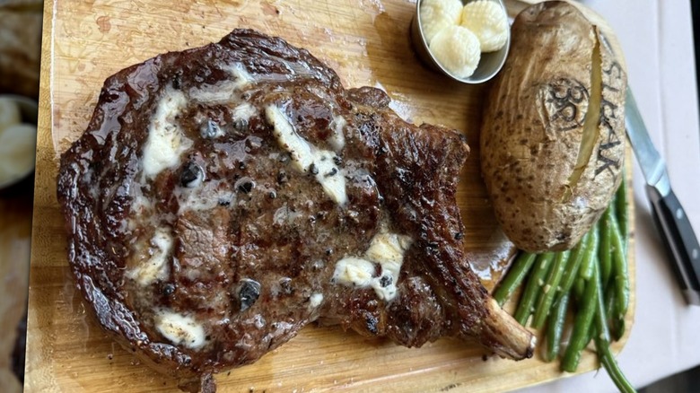 Steak 38 restaurant bone-in prime rib with truffle butter, baked potato, and green beans on wooden cutting board.