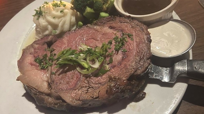 A thick slice of Clyde's prime rib on plate, topped with fresh herbs, served with whipped potatoes, broccoli, creamy horseradish, and au jus.
