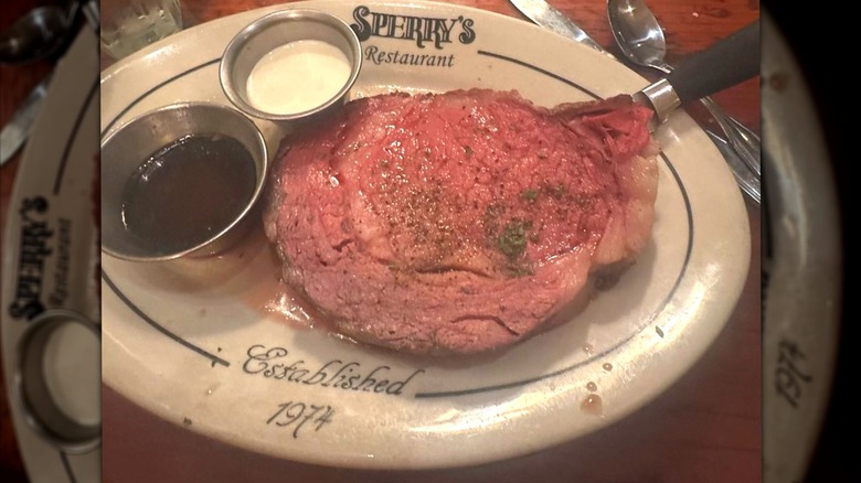 A large slice of Sperry's Restaurant prime rib with cups of au jus and creamy horseradish on plate.