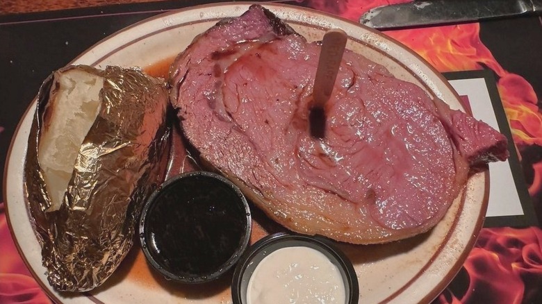 A cut of Timbermine restaurant's prime rib, served with steaming au jus, creamy horseradish, and a gold foiled baked potato on plate.
