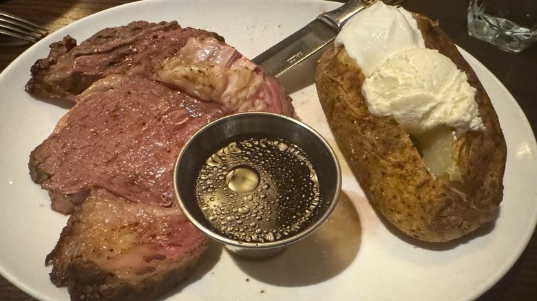 A FireRock Steakhouse prime rib served with a baked potato and cup of au jus on plate.