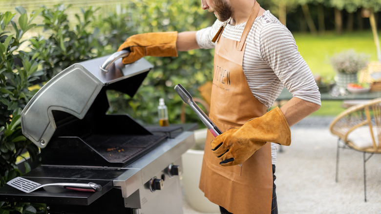 A man with gloves and tongs opening a gas grill