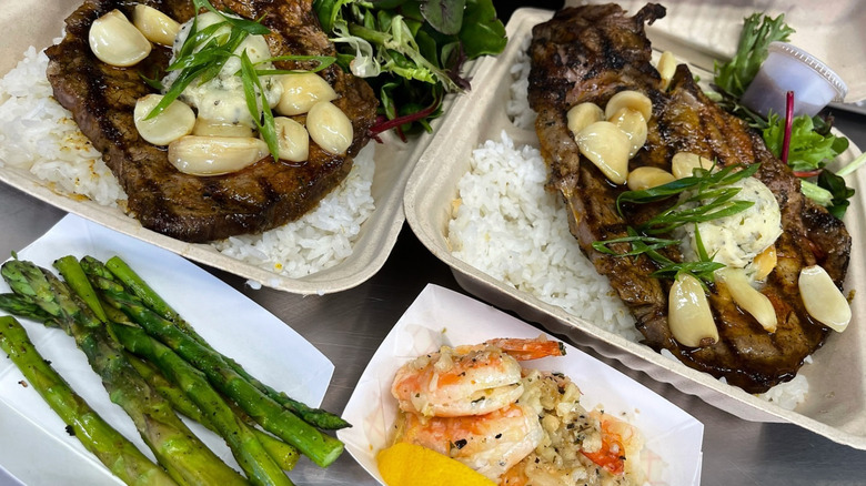 Plates of garlic steak over rice, shrimp, and asparagus in cardboard to-go containers