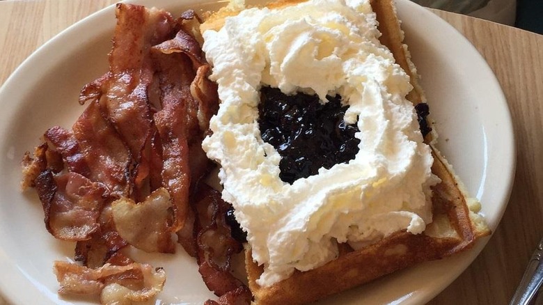 A Belgian waffle topped with blueberry compote and whipped cream, served with bacon