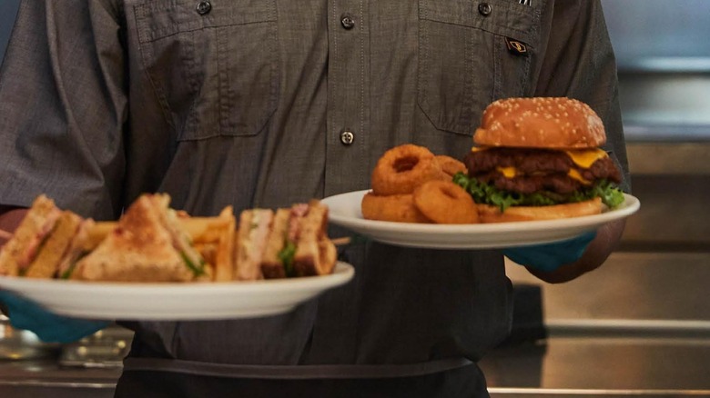 Close-up of a Denny's server carrying two plates, one with a club sandwich, the other with a burger and onion rings