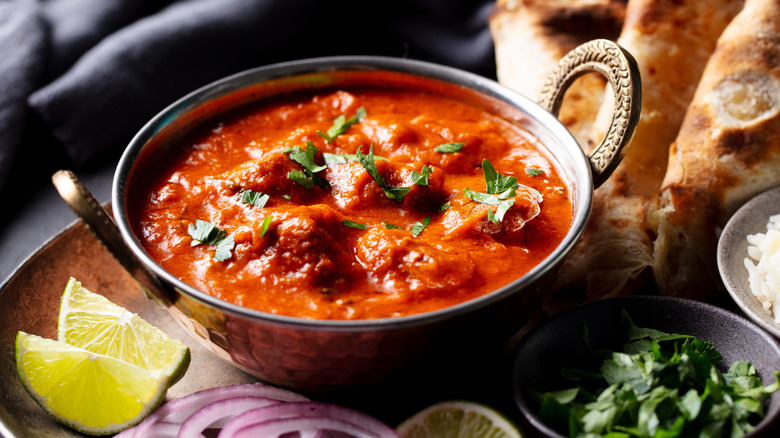 A bowl of Indian curry in a small silver bowl on a plate with other ingredients and sides.