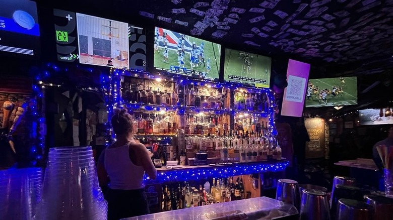 Interior of a dimly-lit bar, facing the barback with bartender on the left, football on three TVs, and dollar bills tacked to the ceiling