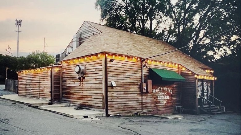 The exterior of a windowless ramshackle building with weathered light brown siding, and a green awning with a "White Water Tavern" sign under it