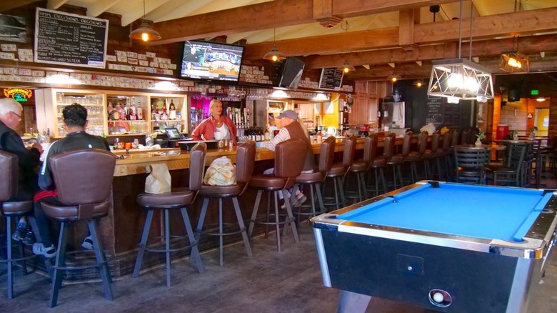 Interior of a rustic bar with wood beams running along the ceiling and a few customers sitting on brown bar stools with a blue felt-topped pool table at the right forefront