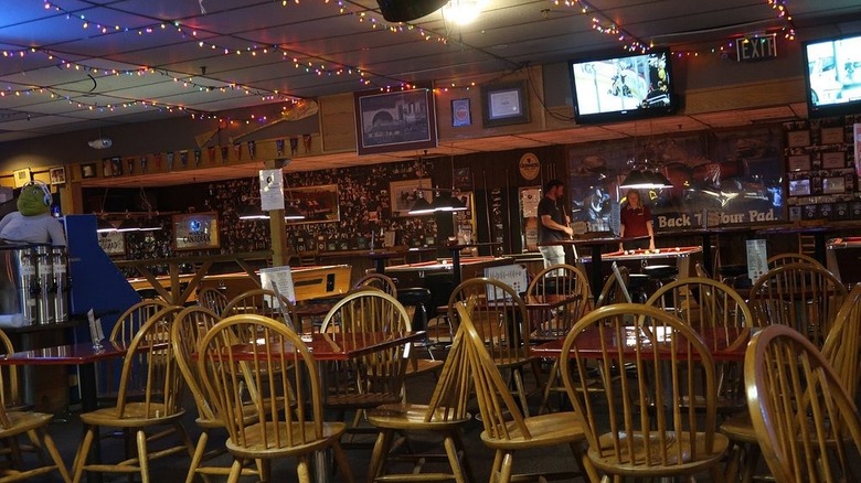 Interior of a nearly empty dimly lit bar with a dropped ceiling strewn with rainbow string lights with two people playing pool in the background
