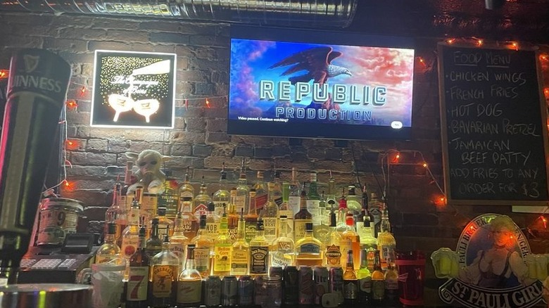 A dimly-lit brick barback with three rows of liquor lined up, red string lights are strewn about and a flat-screen display that reads "Republic Production"