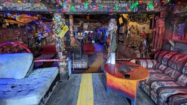 The interior of a bar with mismatched upholstered furniture and dollar bills, string lights, graffiti, and other random objects covering the ceilings, walls, and columns