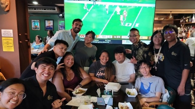 interior of a bar with 12 smiling people posing around a table; some seated, some standing with a big-screen TV in the background