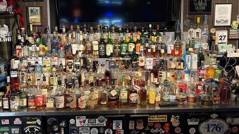 A back bar of about 100 liquor bottles arranged on five shelves; coolers underneath are covered in stickers