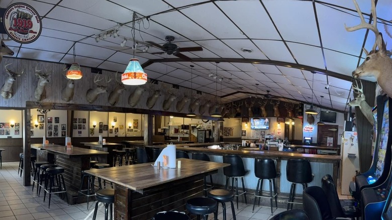 The interior of a bar/restaurant with simple white tile floors, wood tables, and a curved white drop ceiling; taxidermy deer mounts line the walls