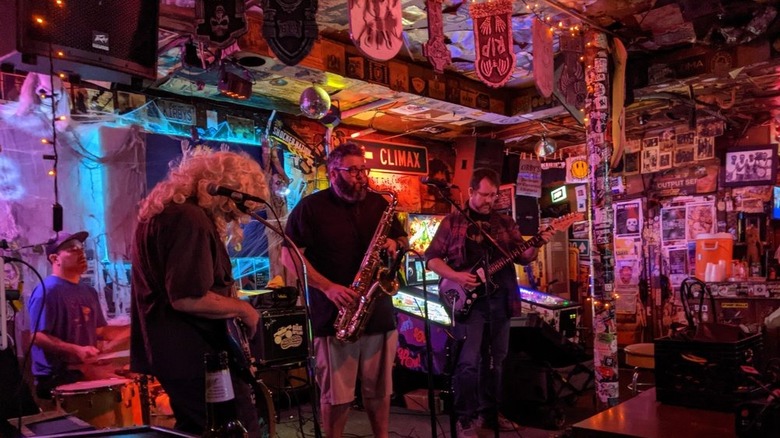 A four-piece band playing in a dimly-lit venue with the walls covered in stickers and posters, and random decor strewn from the ceiling