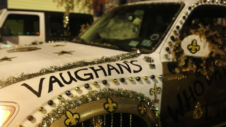Close-up of a white car that is decorated in gold and black baubles and gold tinsel and fleur-de-lis that says "Vaughan's" above the wheel well