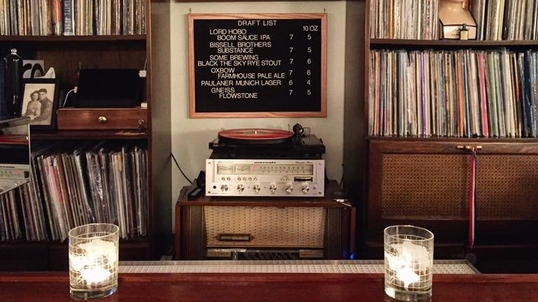 Two ice-filled clear glass tubs sit  on each side of a wooden bar with a turntable in the center flanked by shelves of vinyl records; a letterboard hangs above the turntable with a list of beer on it