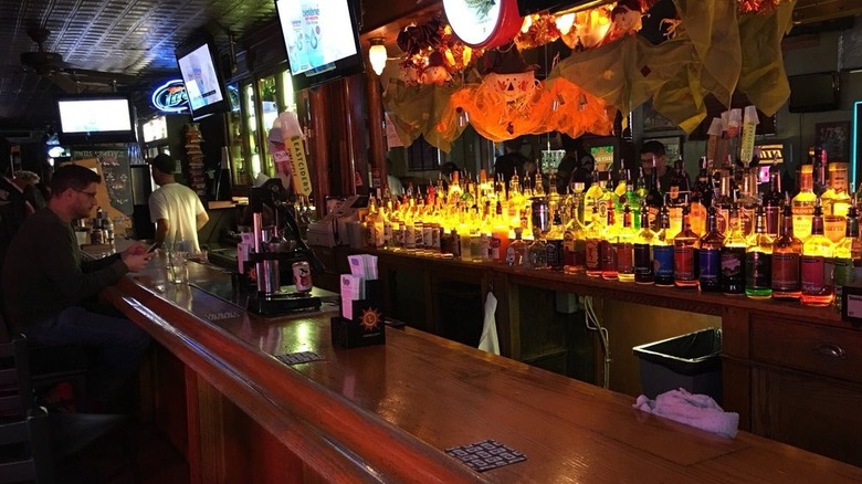 A dimly list bar with one patron sitting on the far left with three shelves of backlit booze bottles on the back bar