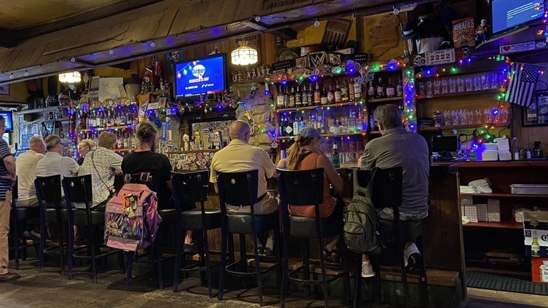 Seven people sitting at a bar, shown from the back; the bar stocked with booze and decorated with rainbow stirng lights and random knick knacks