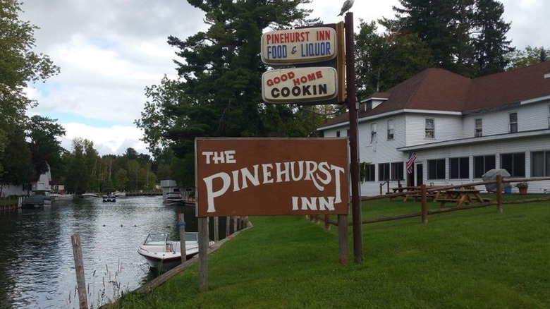 A brown, wood sign that says "The Pinehurst Inn" with two others above it ("Pinehurst Inn Food & Liquor" and "Good Home Cookin" set on a green lawn, with a river to the right and a white two-story building on the left