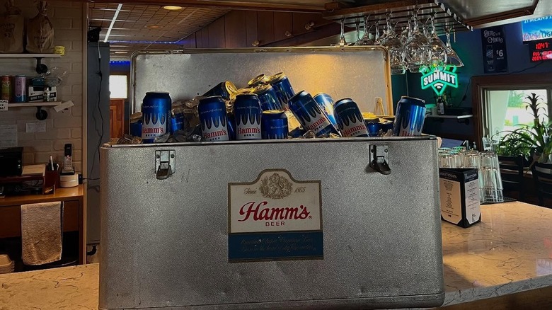 A metal cooler filled with Hamm's beer with a Hamm's beer logo on it sitting on a bar