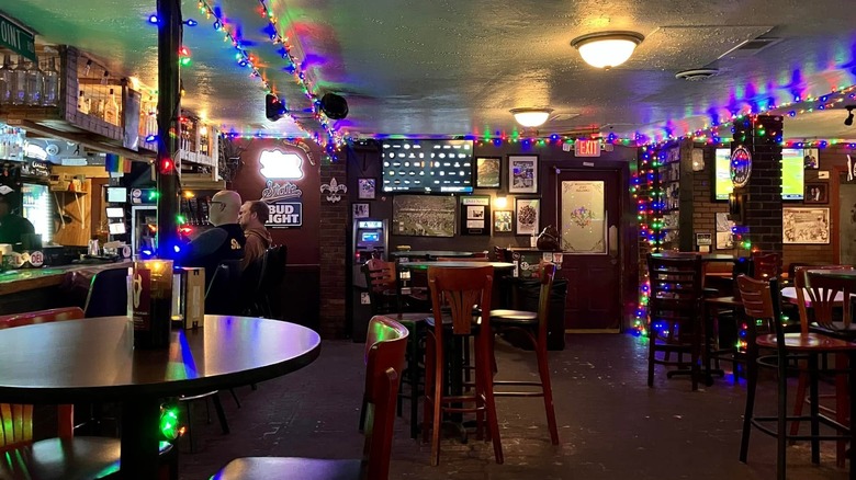The interior of a bar with empty tables lining the right, one in the middle, and another in the left foreground; two men sit at the bar on the left side