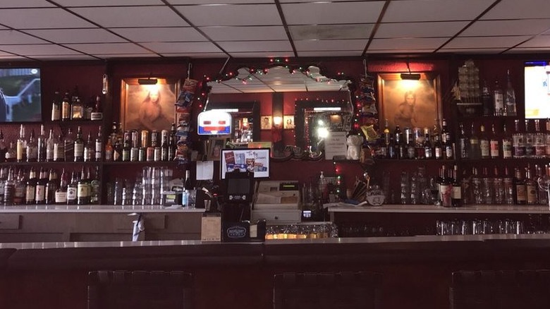 The interior of an empty bar with the center of bar back a mirror flanked by paintings of women is displayed; shelves of booze bottles are on each side