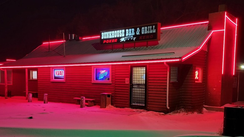 A one-story log-cabin-style building at night lit up by pink neon lights lining its edges; there is snow in the foreground, neon signs in the windows, and a sign on top that read "Bunkhouse Bar & Grill - Poker - Keno