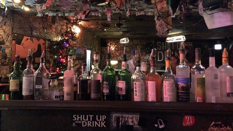 The interior of a bar with liquor bottles lining a shelf and a dimly-lit room with dollar bills tacked to the ceiling and walls along with other random items in the background