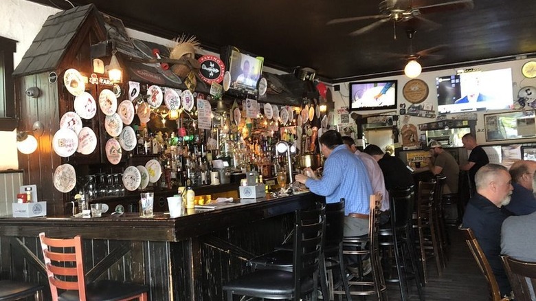 Interior of a small bar with a cluttered dark wood back bar and bar with a few men sitting on wood barstools; a few other men are sitting at a table on the right