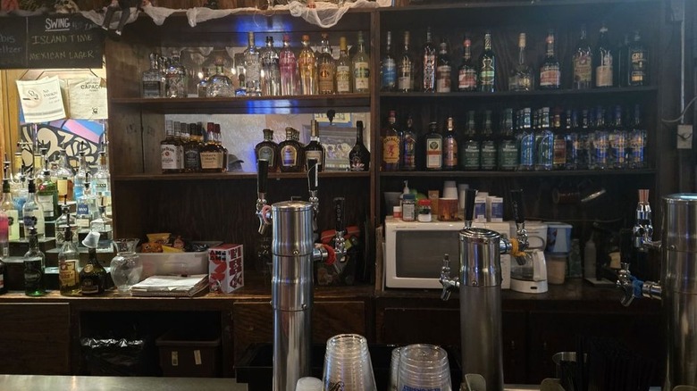 The cluttered wood back bar of a dimly lit bar with two levels of shelves of booze bottles and a microwave and white coffee maker